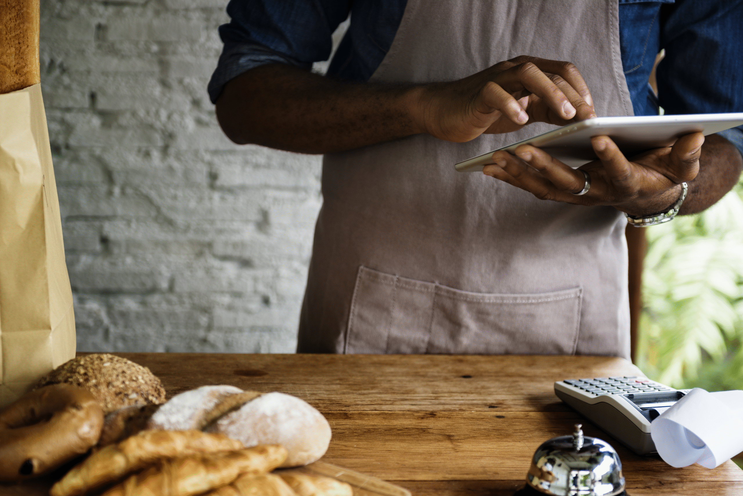 Pessoa usando tablet em padaria para acompanhar métricas para restaurante, com pães e croissants sobre a mesa ao lado de uma maquininha de cartão.