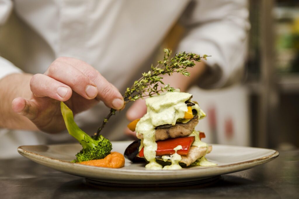 Chef finalizando a apresentação de um prato sofisticado com legumes e molho verde, representando o cuidado e a arte por trás de uma verdadeira experiência gastronômica.