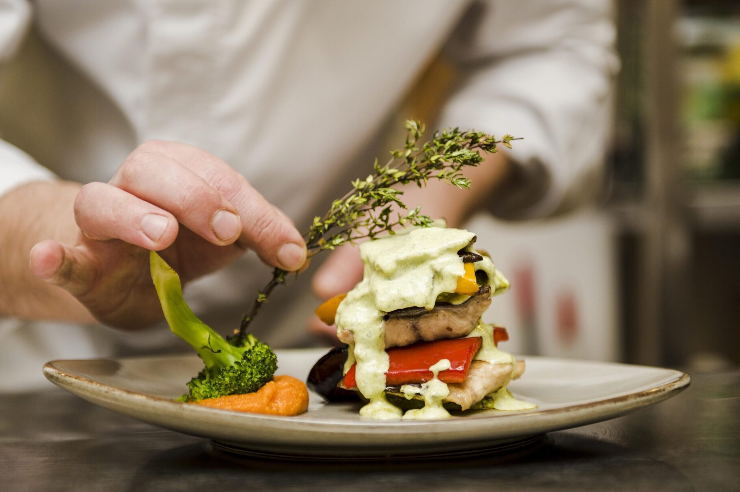 Chef finalizando a apresentação de um prato sofisticado com legumes e molho verde, representando o cuidado e a arte por trás de uma verdadeira experiência gastronômica.