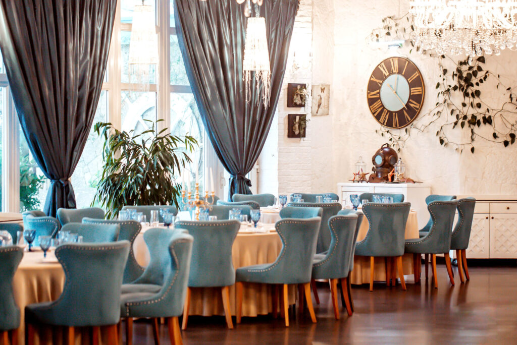 Decoração de restaurantes com ambiente elegante, cadeiras turquesa, lustres de cristal e cortinas escuras, criando um salão sofisticado e acolhedor