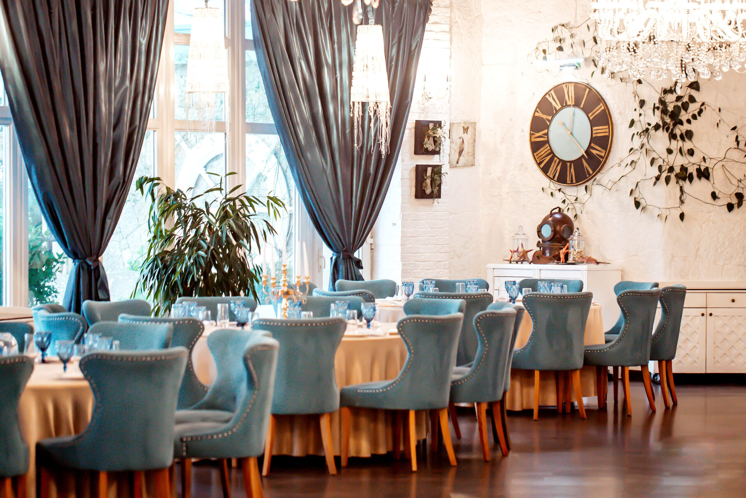 Decoração de restaurantes com ambiente elegante, cadeiras turquesa, lustres de cristal e cortinas escuras, criando um salão sofisticado e acolhedor