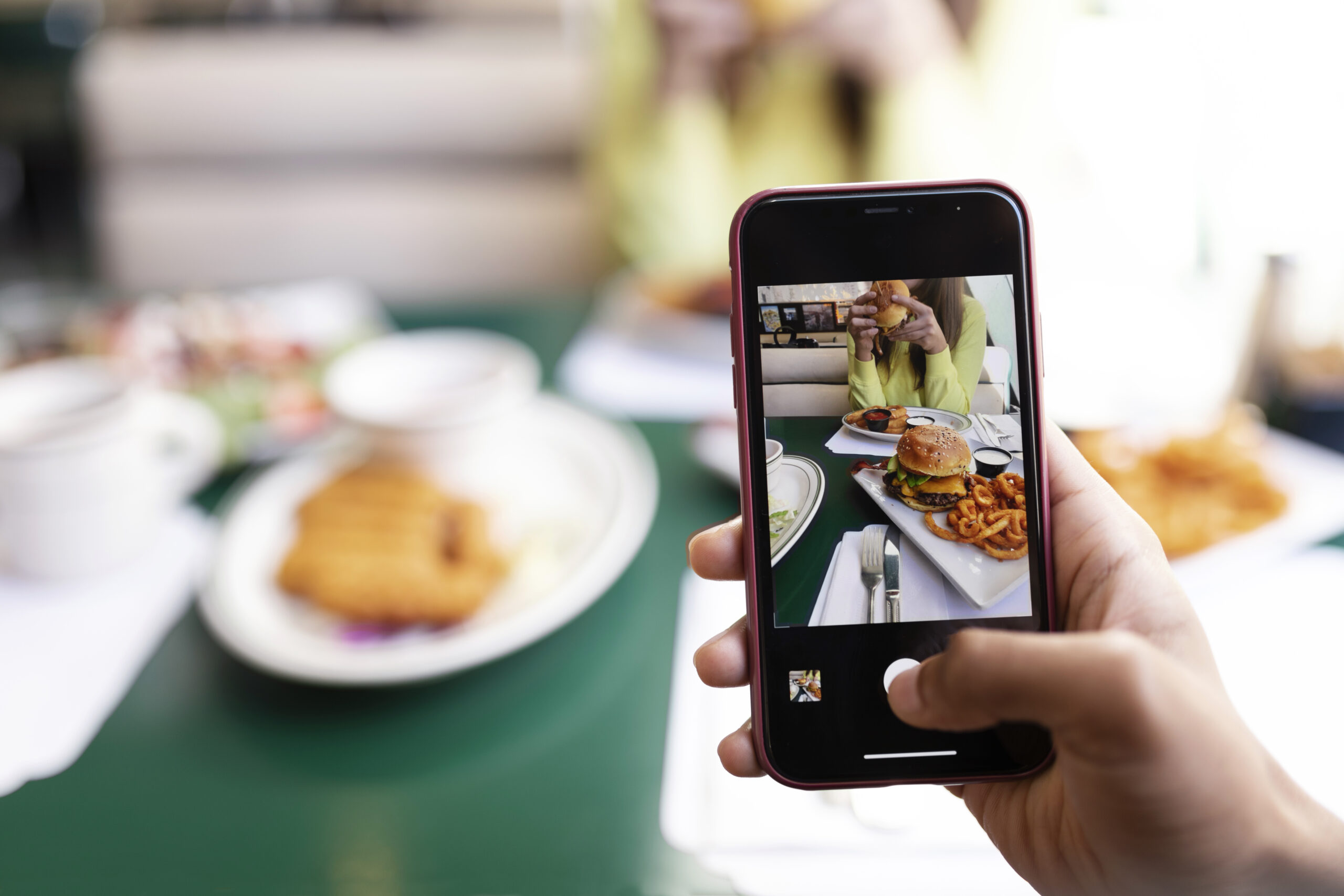 Pessoa fotografando um hambúrguer com o celular em um restaurante, ilustrando como fotos de comida ajudam restaurantes a atrair clientes no Instagram.