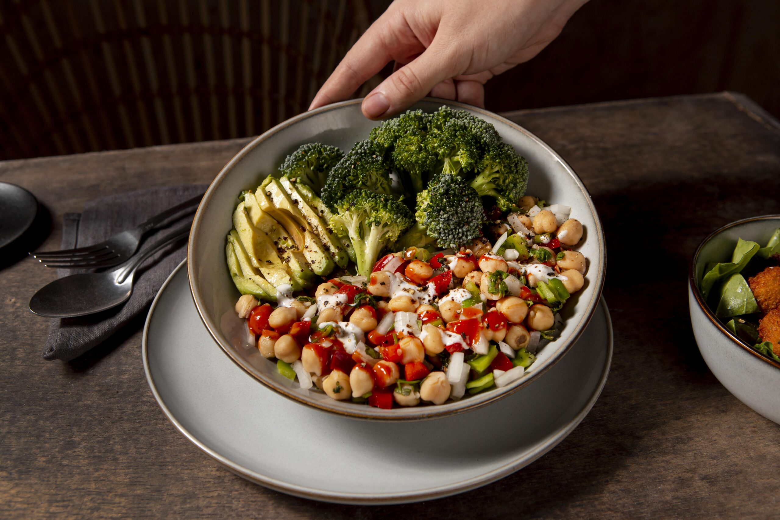 Cardápio inclusivo com bowl saudável de grão-de-bico, brócolis, abacate e legumes frescos servido em prato sobre mesa de madeira.