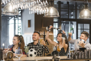 Grupo de amigos em um bar assistindo jogo e torcendo animadamente, com bebidas e bola de futebol sobre o balcão, representando o clima de torcida e socialização durante a Copa do Mundo 2026.