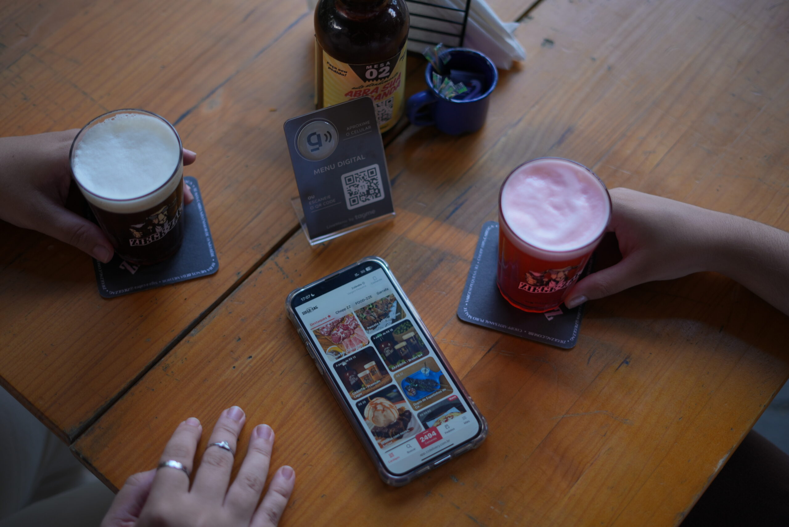 Mesa de restaurante com duas pessoas segurando copos de cerveja, enquanto um celular exibe um cardápio digital para fazer pedidos. Ao centro, um display com QR code para acesso ao menu, reforçando a automação de pedidos no local.