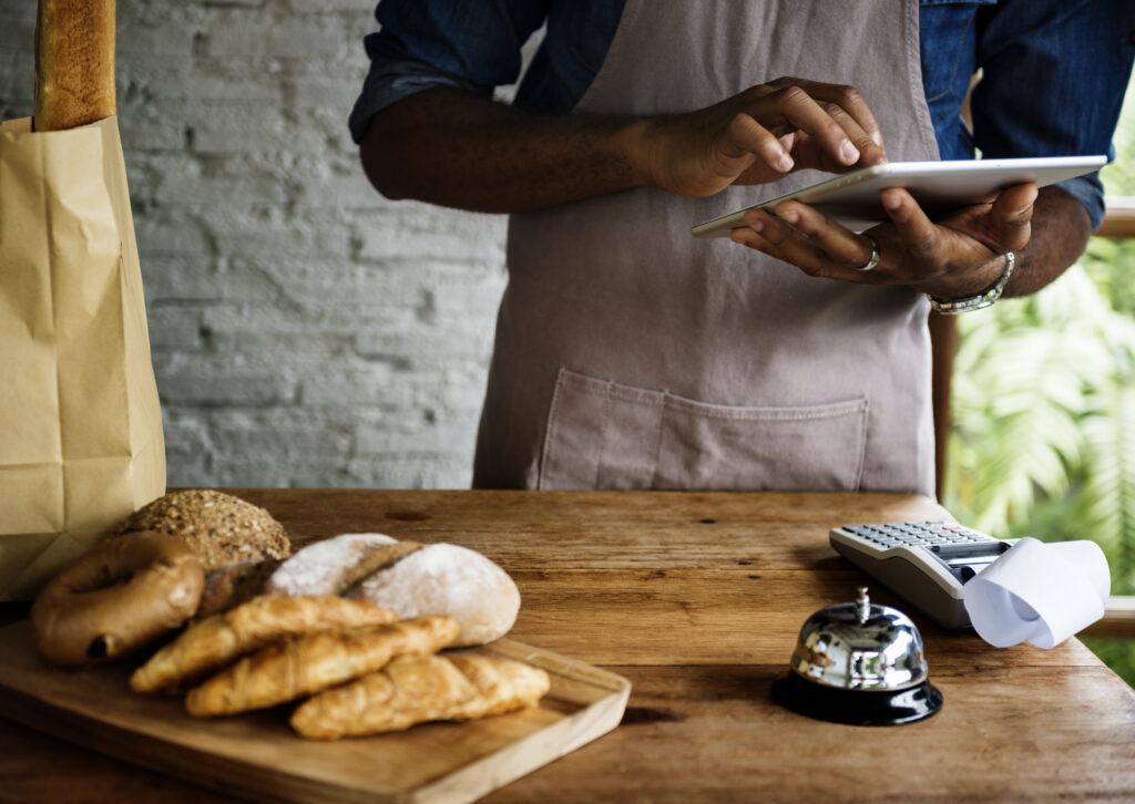 Pessoa usando tablet em padaria para acompanhar métricas para restaurante, com pães e croissants sobre a mesa ao lado de uma maquininha de cartão.