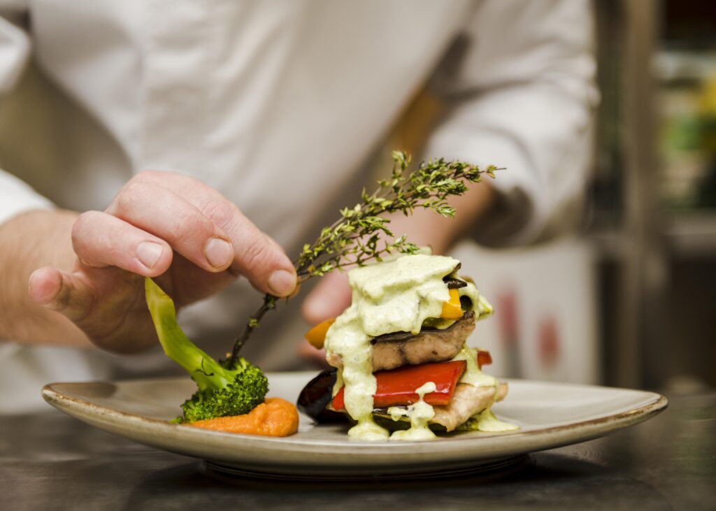Chef finalizando a apresentação de um prato sofisticado com legumes e molho verde, representando o cuidado e a arte por trás de uma verdadeira experiência gastronômica.