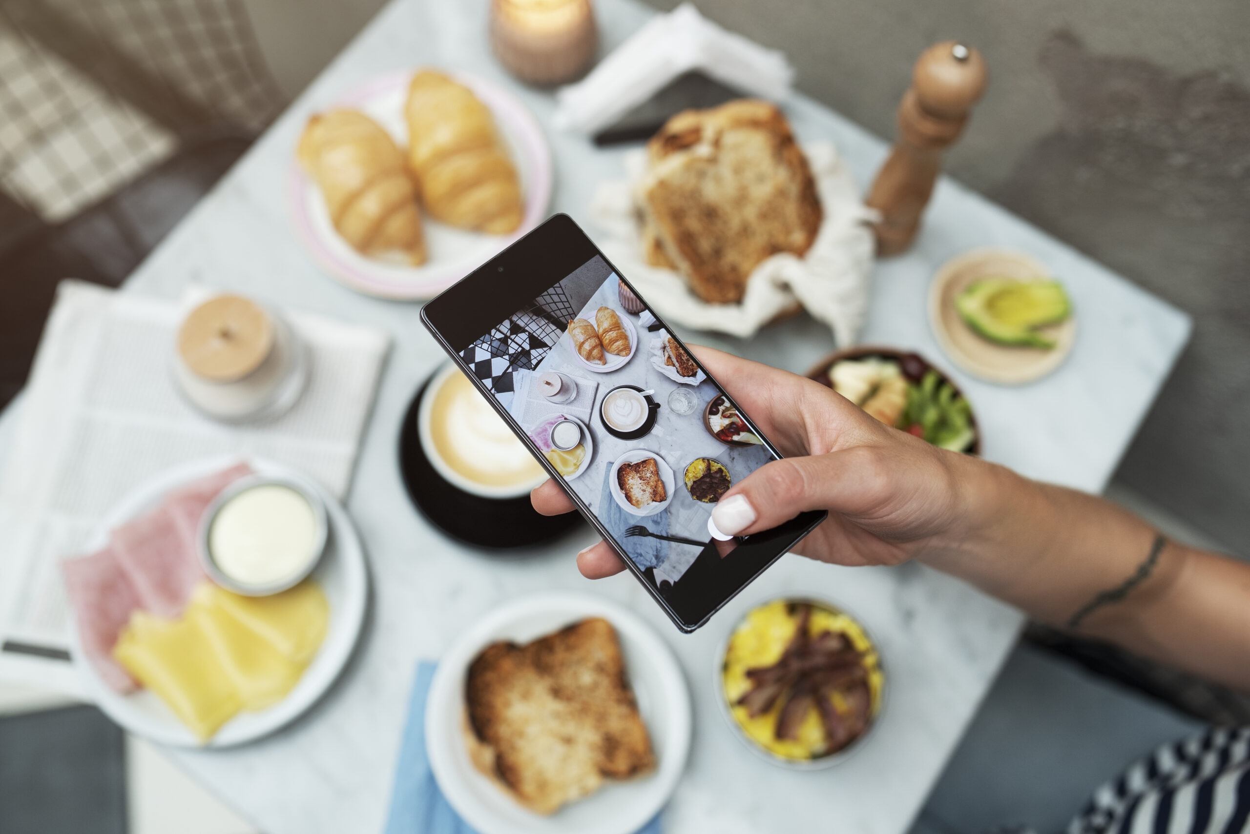 Pessoa fotografando um café da manhã variado para compartilhar como conteúdo para Instagram, com croissants, pães, frios, café e pratos coloridos sobre a mesa.