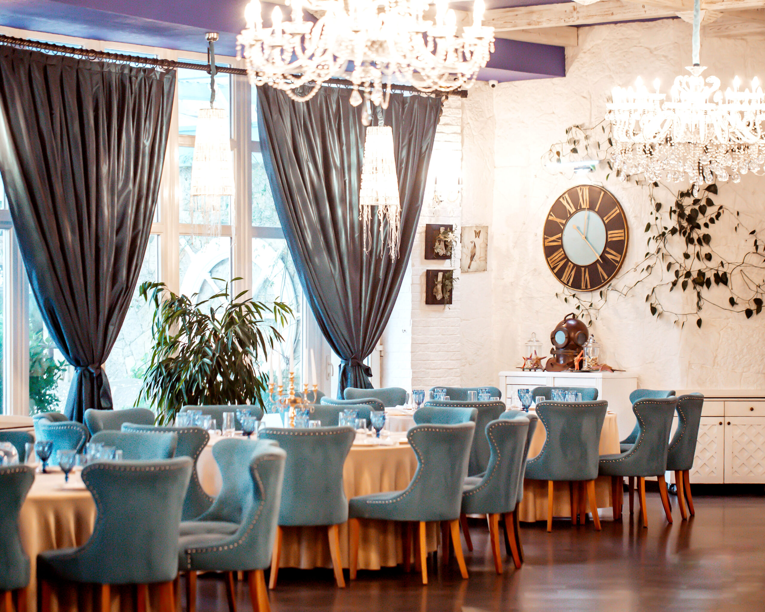 Decoração de restaurantes com ambiente elegante, cadeiras turquesa, lustres de cristal e cortinas escuras, criando um salão sofisticado e acolhedor