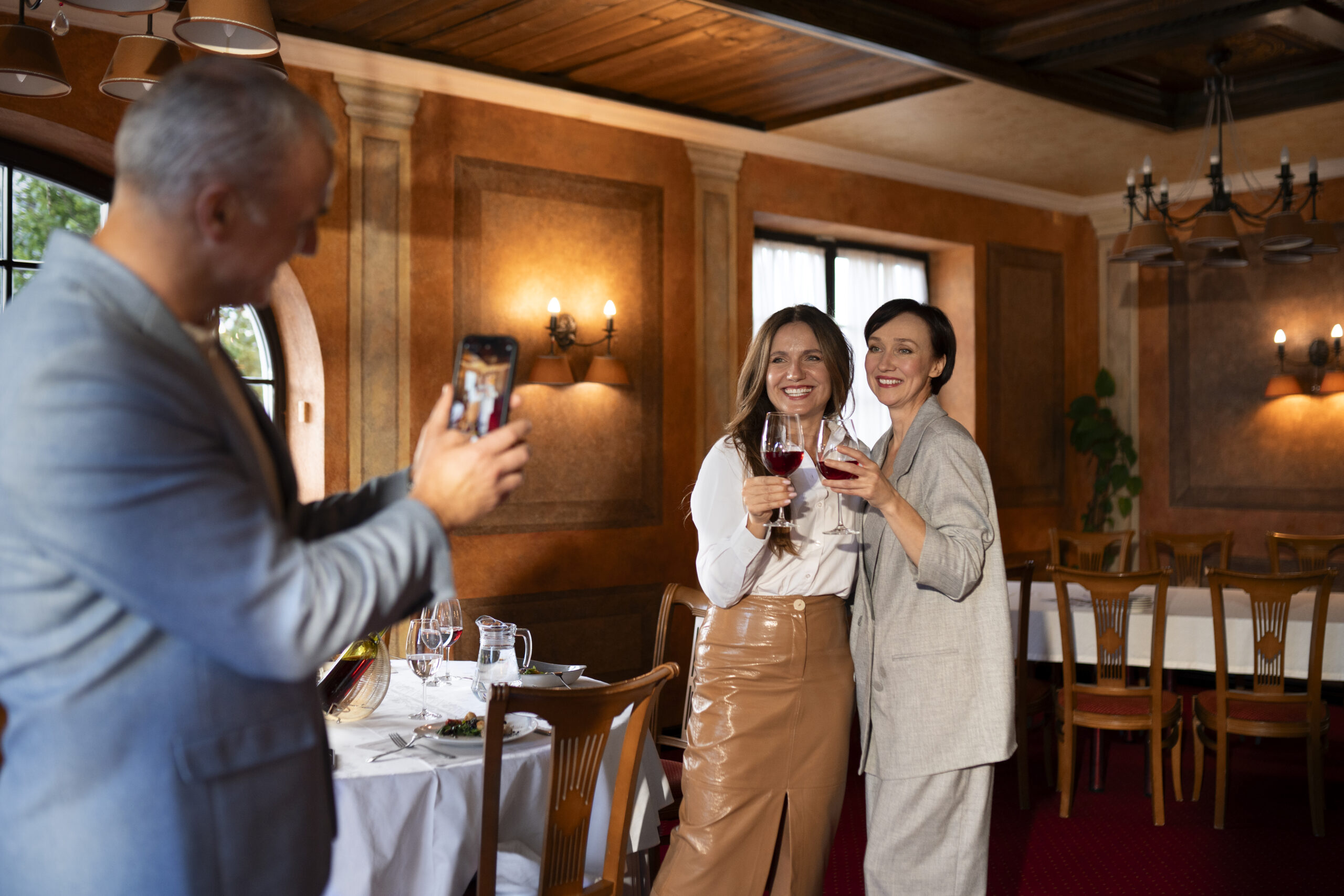 Pessoas brindando com taças de vinho em restaurante elegante, enquanto um homem tira foto da mesa, representando experiências gastronômicas planejadas e reservas antecipadas no calendário 2026.