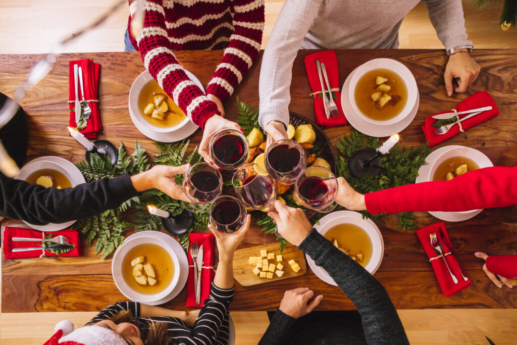Vista superior de um jantar de Natal com pessoas brindando com taças de vinho ao redor de uma mesa decorada, pratos quentes e elementos festivos, representando um momento de confraternização e inspiração para ações de marketing de Natal.