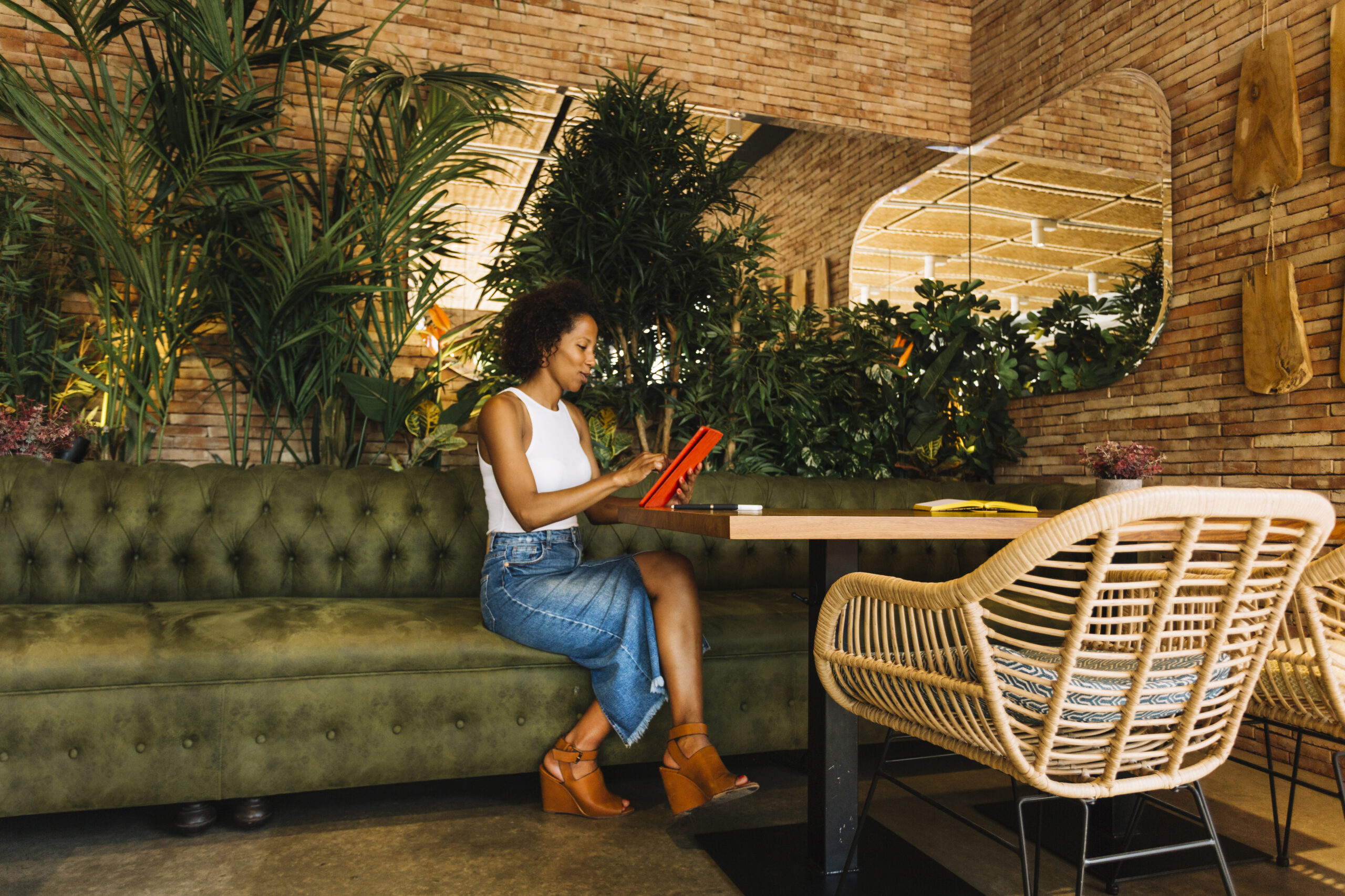 Mulher sentada em um restaurante moderno e aconchegante, usando um tablet à mesa em um ambiente com plantas, madeira e design contemporâneo que representa tendências de restaurante focadas em experiência, tecnologia e conforto.