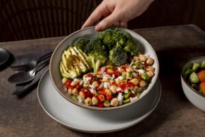 Cardápio inclusivo com bowl saudável de grão-de-bico, brócolis, abacate e legumes frescos servido em prato sobre mesa de madeira.